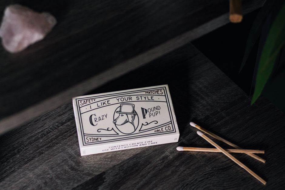 A black and white matchstick safety card featuring playful text and illustrations, placed on a dark wooden surface next to three unlit wooden matches with white tips. The card includes the phrases 'I 