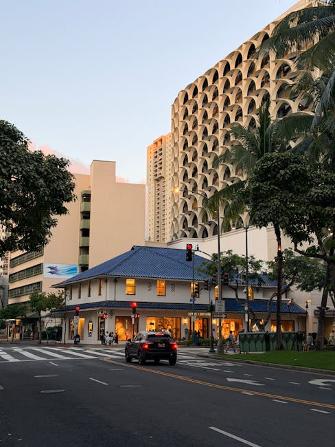 An urban street scene featuring a small, traditional-style building with a blue-tiled roof and warm yellow lighting emanating from its windows, situated at a busy intersection. Surrounding the buildin