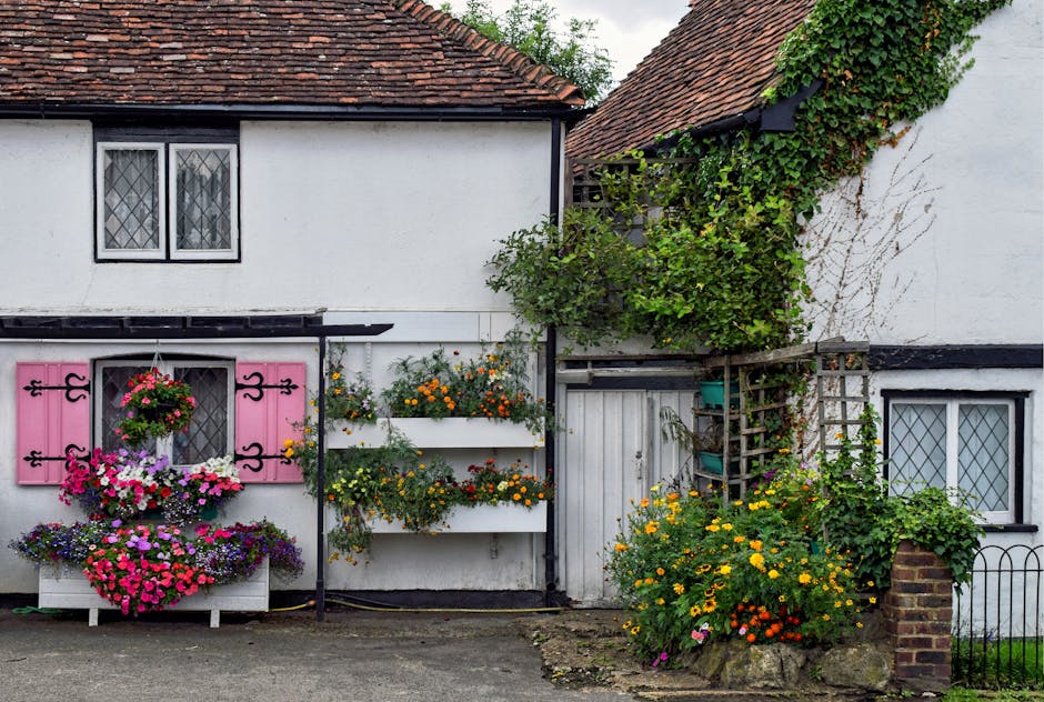 A quaint white cottage with a red tiled roof displays a charming garden in front. The facade features a small window with diamond-patterned panes and pink decorative shutters, complemented by vibrant 