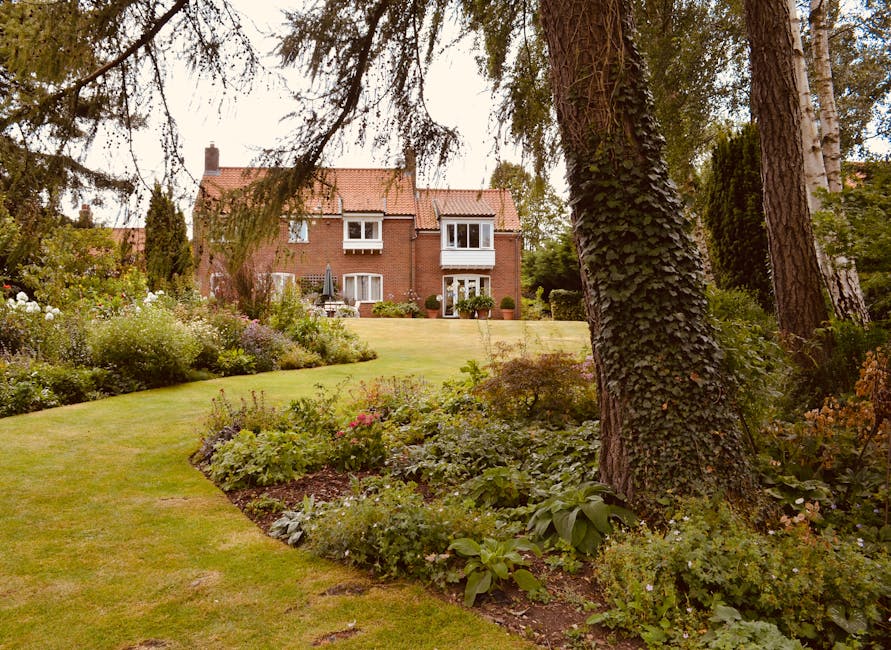 A quaint white cottage with a red tiled roof displays a charming garden in front. The facade features a small window with diamond-patterned panes and pink decorative shutters, complemented by vibrant 